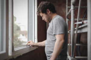 A focused man wearing a casual grey shirt meticulously inspects a white casement window ensuring clarity and shine in a room under renovation.