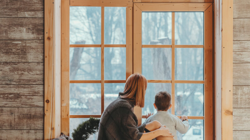A mother and her child enjoying a cozy moment on a wooden windowsill framed by large casement windows overlooking a snowy landscape.