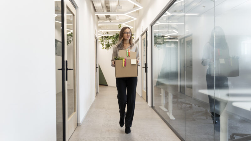 A woman walking through a hallway carrying a box, passing by modern front door glass styles in an office setting.